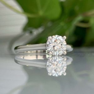 Solitaire diamond engagement ring with a silver band and a round brilliant-cut diamond, resting on a reflective surface.