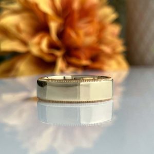 Close-up of a gold ring with milgrain edge resting on a reflective surface, with a blurred autumn bouquet in the background
