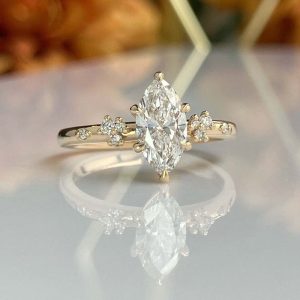 Gold engagement ring with a marquise-cut center diamond and small side stones, reflected on a glass surface.