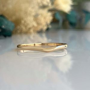 Gold wave-shaped ring resting on a reflective surface with a blurred floral background
