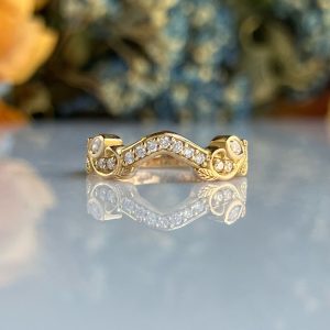 Gold ring with a curved row of diamonds, resting on a reflective surface with a blurred floral background.
