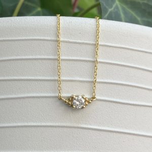 Gold chain necklace with a central round diamond held by six bead-like prongs against a textured light background, minimalist design.