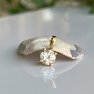 Gold pendant with a round-cut diamond resting on a light stone, reflecting on a glossy surface