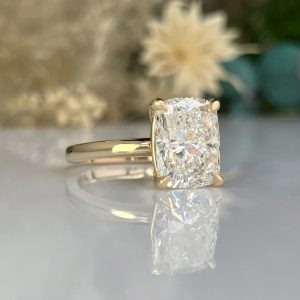Gold engagement ring with a cushion-cut diamond, reflected on a glassy surface.
