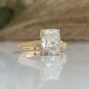 Gold engagement ring with a cushion-cut diamond in a prong setting and ornate engraving along the band, reflected on a glass surface.