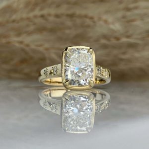 Gold band cushion-cut diamond engagement ring with smaller diamonds along the sides and a mirrored reflection on a smooth surface.