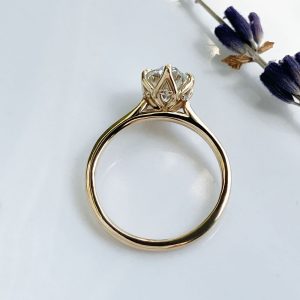 Gold engagement ring with a crown-like setting holding a central diamond on a white background with a lavender sprig nearby, close-up detail