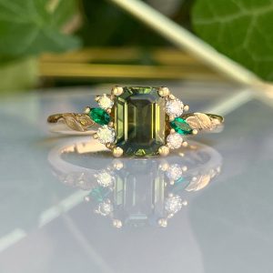 Gold band ring with an emerald-cut green center stone, surrounded by diamonds and green leaf accents on each side.