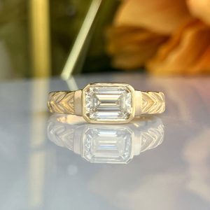 Gold ring with an emerald-cut center diamond and chevron-patterned band, reflected on a glossy surface.