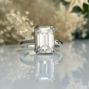 Emerald-cut diamond engagement ring on a silver band, with a subtle floral backdrop.