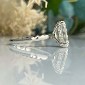 Silver engagement ring with a rectangular diamond and small side stones set in a V-shaped bezel on a mirrored surface.