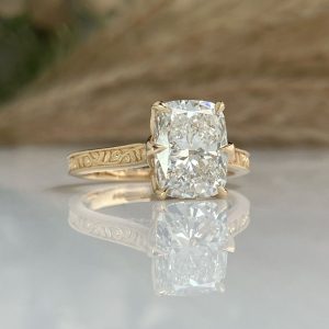 Gold engagement ring with an oversized cushion-cut diamond and engraved band, reflected on a surface.