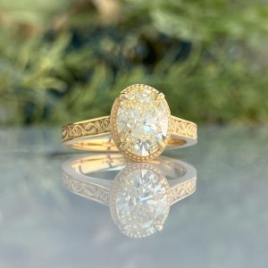 Gold engagement ring with an oval-cut diamond and decorative engraved band, reflected on a glossy surface.