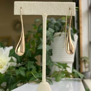 Gold teardrop earrings hanging from a white display stand against a leafy green background.