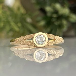 Gold ring with a bezel-set round diamond and leaf-patterned band, reflected on a glossy surface.