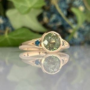 Rose gold ring with a round green gemstone and small blue accent stones, reflected on a glossy surface.