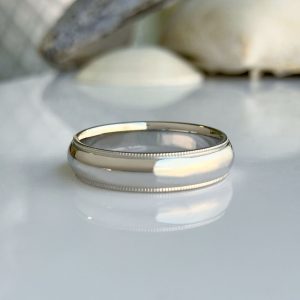 Polished silver wedding band with a beaded edge resting on a reflective white surface, blurred stone background.