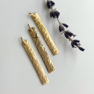 Three gold hammered rectangular bar earrings with hook tops, arranged on a light surface with dried lavender nearby, showcasing a minimalist pendant style.