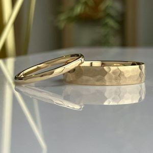 Two gold rings resting on a reflective surface: one smooth, polished band and one with a hammered texture.