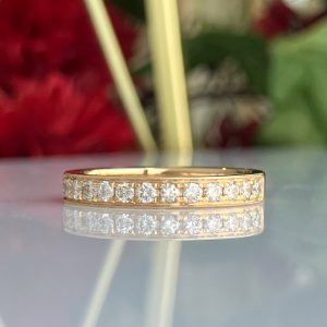 Close-up of a textured gold ring with diamond-like stones, resting on a reflective surface with pale vertical rods crossing in the foreground.