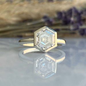 Silver ring featuring a hexagonal-cut diamond centerpiece on a reflective surface, with a blurred background.