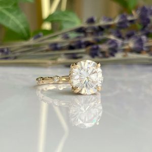 Gold engagement ring with a large round-cut center diamond and small side stones on the band, reflected on a glossy surface with a blurred floral background.