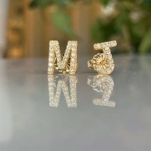 Gold initial jewelry letters M and J encrusted with diamonds, resting on a reflective surface.
