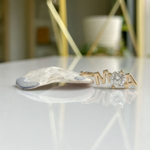 Gold 'MIA' pendant with a diamond where the 'I' would be, resting on a glossy white surface.