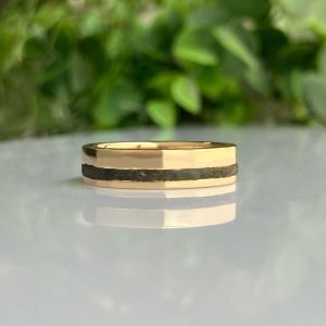 Gold ring with a black central inlay rests on a glossy surface, greenery blurred in the background.