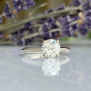 Solitaire diamond engagement ring with a round-cut gem on a silver band, reflected on a glossy surface with lavender flowers in the background