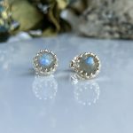 Pair of silver stud earrings with round blue iridescent gemstones set in a scalloped bezel, reflecting light on a white surface.