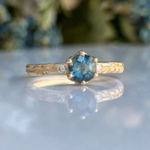 Gold ring with a blue cushion-cut center stone and small side diamonds, on a reflective surface.
