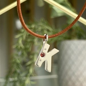 Silver letter 'K' pendant hanging on a brown cord necklace, featuring red gemstone and small clear stones.