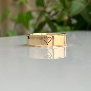 Gold wedding band with a heart outline engraving, resting on a reflective surface.