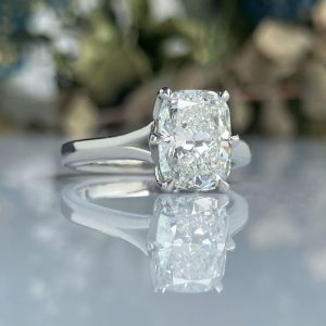 Engagement ring with a cushion-cut diamond in a silver band, reflected on a glossy surface.
