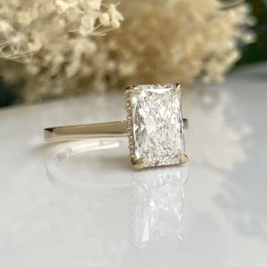 Emerald-cut diamond ring with small side stones on a gold band, on a reflective white surface.