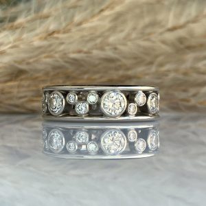 Close-up of a metallic jewelry piece—row of clear, round-cut stones set in a band, resting on a reflective surface with a blurred background