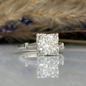 Princess-cut diamond engagement ring with a silver band, shown on a reflective surface.”,