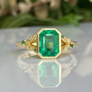 Gold ring with a large emerald-cut green gemstone and leaf-patterned band, reflected on a glossy surface.