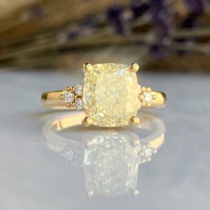 Gold cushion-cut yellow diamond engagement ring with small side diamonds on a four-prong setting, reflected on a glossy surface.