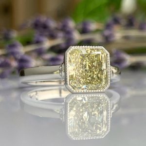 Silver ring with a large pale yellow cushion-cut diamond on a reflective surface, blurred purple flowers in the background