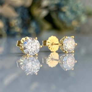 Gold round-cut diamond stud earrings set in yellow gold, placed on a reflective surface with mirrored image.