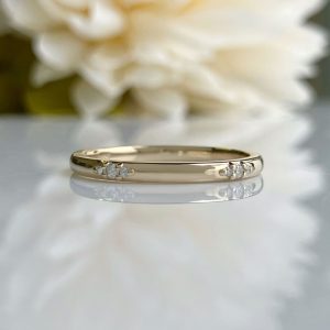 Gold yellow-gold band with small diamonds on both sides, resting on a reflective surface with soft white flowers blurred in the background.