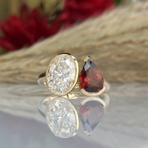 Gold ring set featuring a large oval diamond and a red pear-shaped gemstone on a reflective surface.