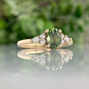 Gold ring with an oval green center stone and diamond accents on the band, reflected on a glossy surface.
