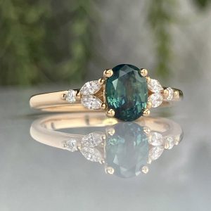 Gold ring with an oval green gemstone set in four-prong halo, flanked by diamond side stones on a reflective surface.