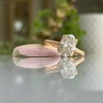 Rose gold engagement ring with a cushion-cut diamond, reflected on a glass surface beside a pink petal.