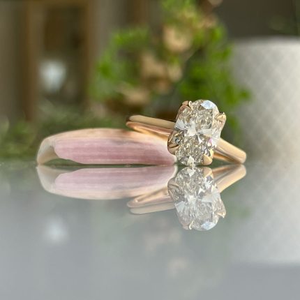 Rose gold engagement ring with a cushion-cut diamond, reflected on a glass surface beside a pink petal.