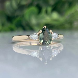 Gold ring with a pear-shaped dark green center gemstone and two side diamonds, reflected on a glass surface.