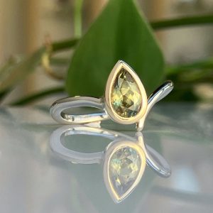 Silver ring with a pear-shaped pale green gemstone, set in a smooth bezel, shown on a reflective surface with blurred green background.